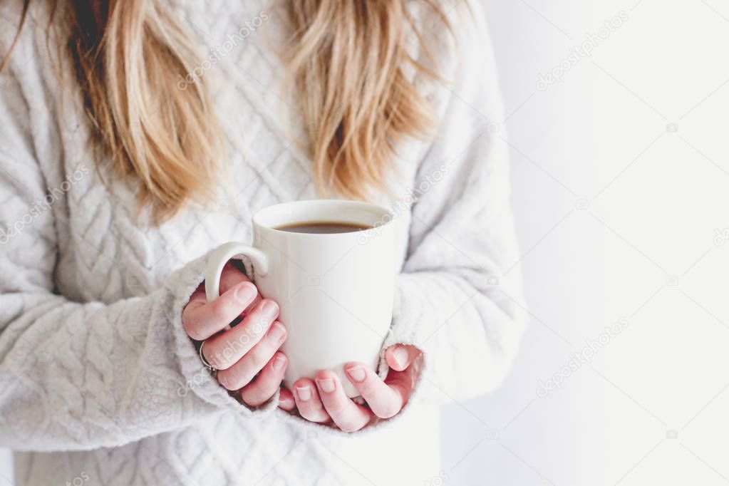 A midsection of a blonde haired girl holding up a white cup on an isolated white background in a cosy lifestyle image.