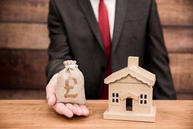 Emlakçı konsepti ev fiyatlarını, evleri, ipotekleri ve ev sigortasını gösteriyor. Yeni evin yanında bir çanta dolusu para tutan bir emlakçı var.