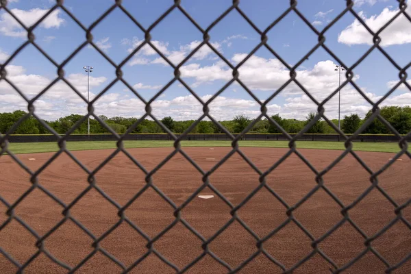 美しい春の日にホームプレートの後ろからチェーンリンクフェンスのダイヤモンド模様のグリッドを通って野球場を見てください — ストック写真