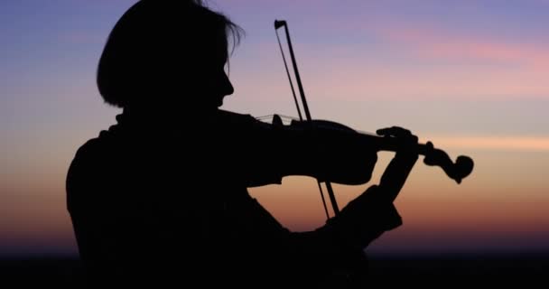 Silhouet meisje violist spelen van de viool bij zonsondergang hemelachtergrond. Slow motion 4k. — Stockvideo