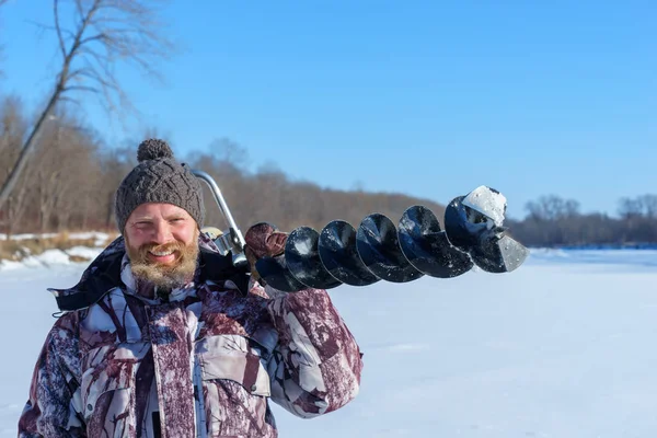 Bearded man is drilling ice hole by automatic moto boer for winter fishing at sunny day under blue sky — Stock Photo, Image