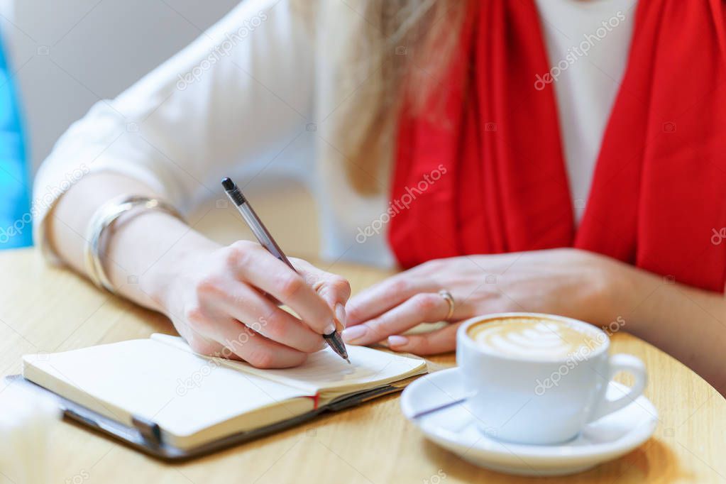 European woman with red scarf is writing by pen somthing in the notepad near white cup of coffee on table