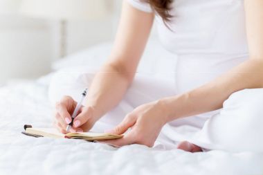 Bir Avrupa hamile kadın güneşli gün ışık Oda beyaz yatak üzerinde oturan pen tarafından not defterinde bazı fikir veya thouths yazar.