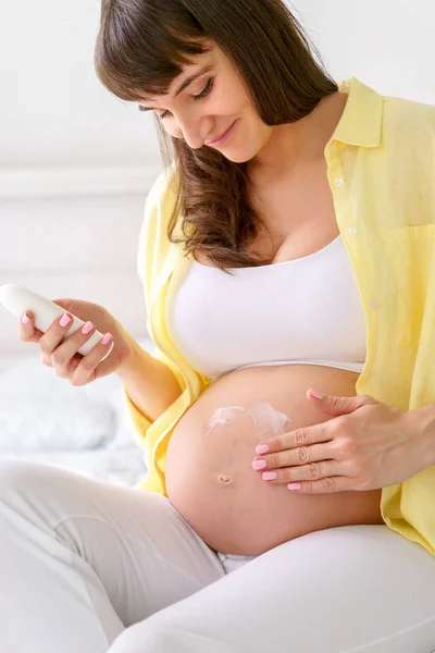 Una mujer embarazada europea con sonrisa está usando crema hidratante en su barriga para evitar las estrías del embarazo mientras se sienta en una cama blanca en la habitación de luz en el día soleado — Foto de Stock