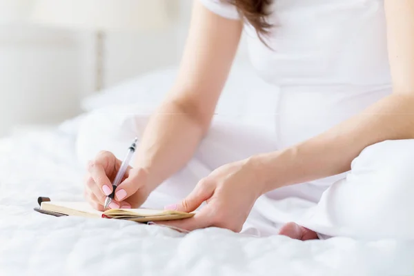 Una mujer embarazada europea escribe alguna idea o pensamientos en el cuaderno de notas por pluma sentado en la cama blanca de la sala de luz en el día soleado . — Foto de Stock