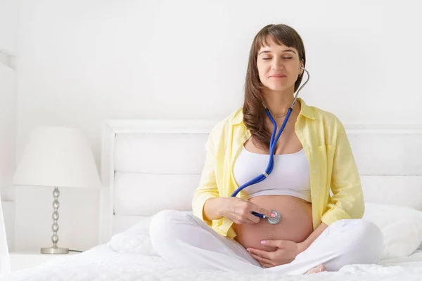 Una mujer embarazada europea con sonrisa está escuchando latidos del corazón del niño por nacer dentro de su barriga por estetoscopio mientras se sienta en la cama blanca en el dormitorio de luz en el día soleado — Foto de Stock