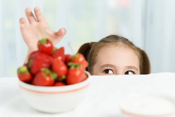 Joven linda sonriente niña europea está tratando de robar maduro jucy fresa de plato de muchas bayas, mientras que ella sentada debajo de la mesa blanca — Foto de Stock