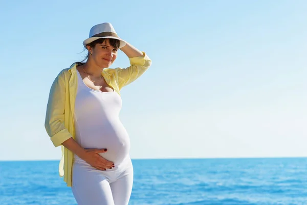 Joven mujer embarazada europea atractiva en camisa de color amarillo y sombrero blanco se queda en la orilla del tranquilo mar azul y la celebración de la barriga a mano derecha bajo el cielo despejado en el día soleado — Foto de Stock