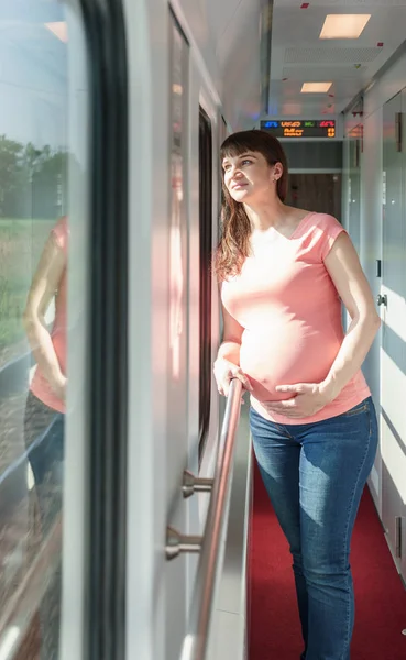 Pregnant european woman is staying and holding her own tummy close to train window while she traveling by rail way and holding handrail inside the carriage