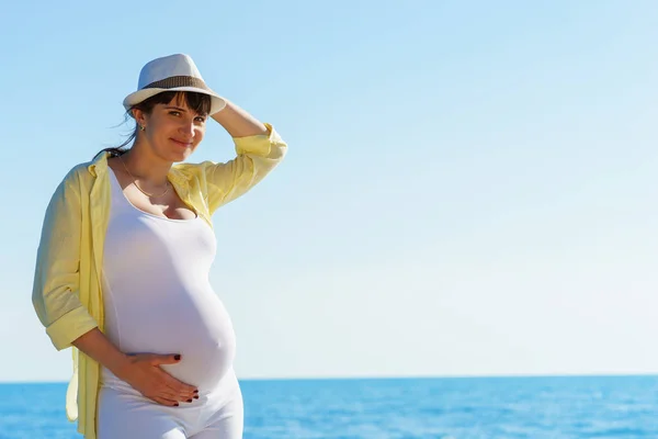 Joven mujer embarazada europea atractiva en camisa de color amarillo y sombrero blanco se queda en la orilla del tranquilo mar azul y la celebración de la barriga a mano derecha bajo el cielo despejado en el día soleado — Foto de Stock