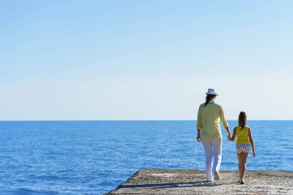 Europese zwangere moeder en haar dochtertje lopen op de golfbreker aan de blauwe zee onder de heldere hemel hand in hand voor elkaar — Stockfoto