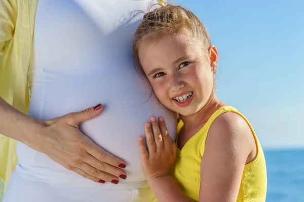 Joven europea hija es tocando la barriga de su embarazada atractiva mamá cerca del mar bajo el cielo azul — Foto de Stock