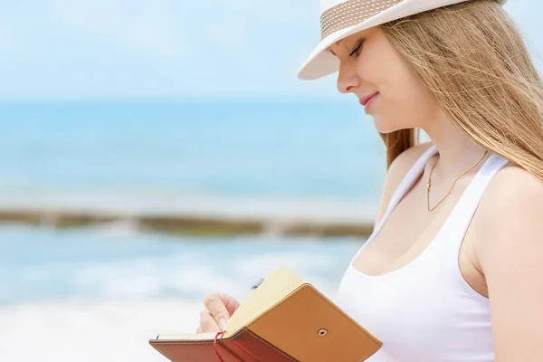 Jeune fille attrayante avec un sourire gentil écrit une idée ou une lettre dans son carnet de notes par stylo sur fond de mer bleue — Photo