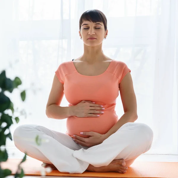 Una mujer embarazada europea con camiseta rosa hace ejercicios de yoga para cuidar de su salud y del feto en un dormitorio luminoso durante el día — Foto de Stock