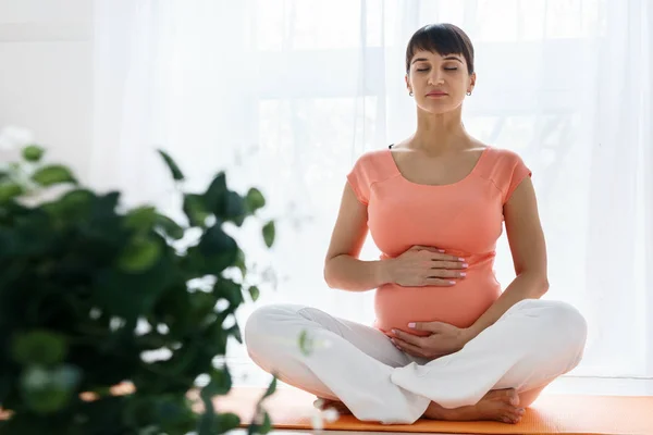 Una mujer embarazada europea con camiseta rosa hace ejercicios de yoga para cuidar de su salud y del feto en un dormitorio luminoso durante el día — Foto de Stock