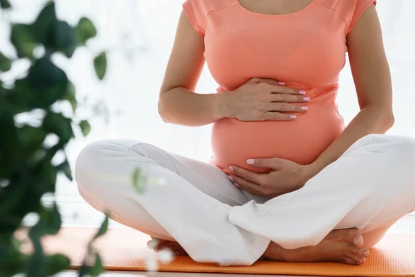 Una mujer embarazada europea con camiseta rosa hace ejercicios de yoga para cuidar de su salud y del feto en un dormitorio luminoso durante el día — Foto de Stock