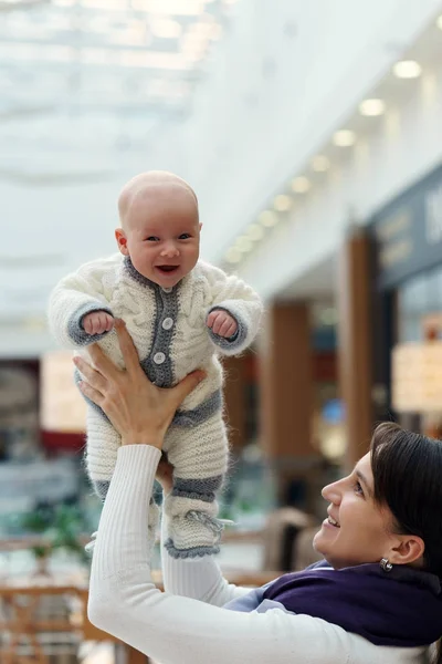 Young caucasian mom is playing with her cute jolly infant son, throwing him up and catching again at public place — Stock Photo, Image