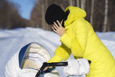 Sarı ceketli beyaz anne beyaz bebek arabası ile kış güneşli gün orman arasında kar yolda yürüyor