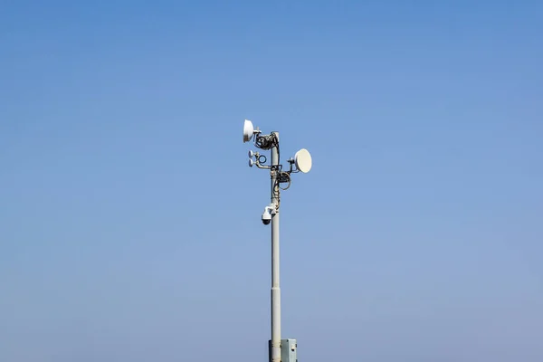 Security and satellite system with a surveillance camera, antenna and communications dishes on a pole against a sunny blue sky