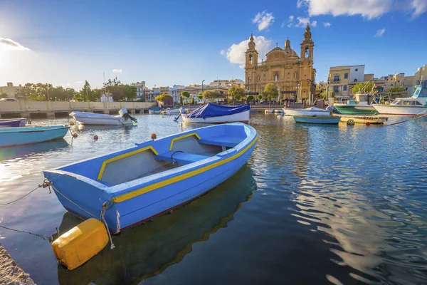 Мсида, Мальта - Синяя традиционная рыбацкая лодка со знаменитой приходской церковью Мсида на заднем плане в летний день с голубым небом и облаками — стоковое фото