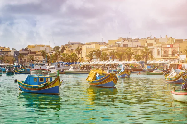 Marsaxlokk, Malta เรือประมงมอลต์ลูซุสีสันแบบดั้งเดิมที่หมู่บ้านเก่าของ Marsaxlokk ด้วยน้ําทะเลเทอร์ควอยส์และต้นปาล์มในวันฤดูร้อน — ภาพถ่ายสต็อก