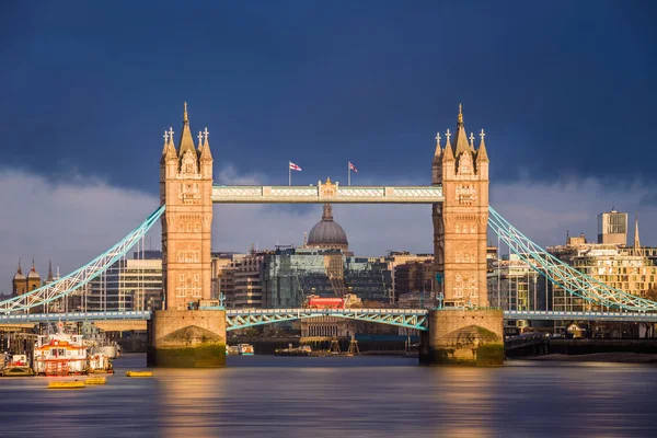 London, England - die weltberühmte Tower Bridge bei goldenem Sonnenaufgang mit rotem Doppeldeckerbus. St. Pauls Kathedrale und dramatischer dunkler Himmel im Hintergrund — Stockfoto