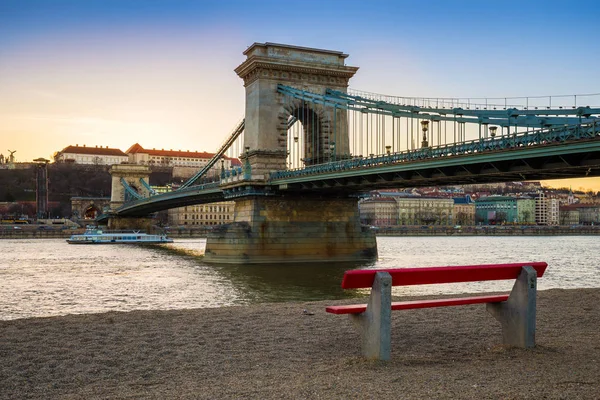 Будапешт, Венгрия - Цепной мост Фетхие с традиционной красной скамьей и прогулочным судном на реке Дунай на закате. Ясное голубое небо — стоковое фото