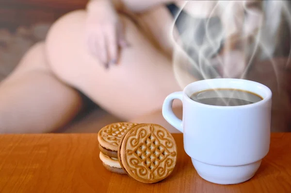 A cup of hot coffee and round cookies with a silhouette of a sexy girl in black underwear in the background on a bed. Focus foreground priority. Small white coffee cup with steam and brown biscuits