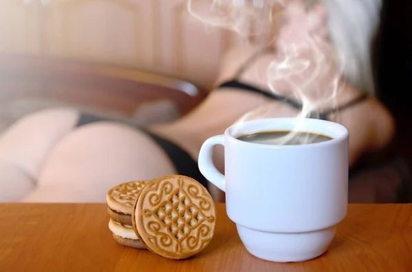 A cup of hot coffee and round cookies with a silhouette of a sexy girl in black underwear in the background on a bed. Focus foreground priority. Small white coffee cup with steam and brown biscuits