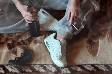 A girl sitting on the couch, smoking a cigarette, drinking beer and playing on a game console in a smoky interior. The concept of wrong unhealthy lifestyles and bad habits clipart