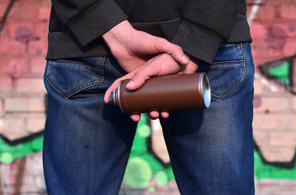 Вид сзади на тело молодого парня в синих грязных джинсах и серый свитер с брызгами краски в руке на фоне старой кирпичной стены, нарисованной граффити
