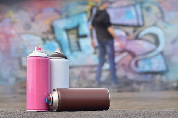 A photography of a certain number of paint cans against the background of the space with the wall on which the young guy draws a large graffiti drawing. Modern art of drawing walls in graffiti
