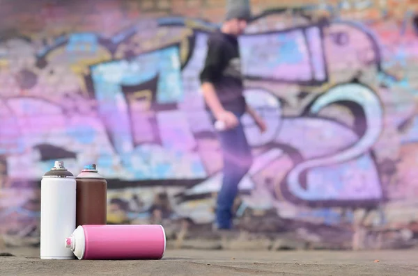 A photography of a certain number of paint cans against the background of the space with the wall on which the young guy draws a large graffiti drawing. Modern art of drawing walls in graffiti
