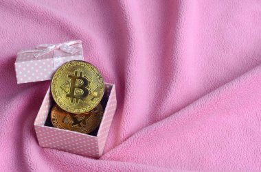 Küçük pembe hediye kutusu yumuşak ve kabarık hafif pembe Polar kumaş çok sayıda kabartma kıvrımları ile yapılmış bir battaniye üzerinde küçük bir yay ile altın bitcoin yatıyor