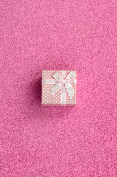 A small gift box in pink with a small bow lies on a blanket of soft and furry light pink fleece fabric. Packing for a gift to your lovely girlfriend