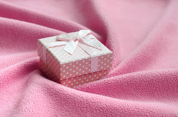 A small gift box in pink with a small bow lies on a blanket of soft and furry light pink fleece fabric with a lot of relief folds. Packing for a gift to your lovely girlfriend