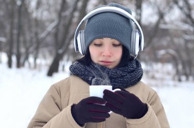 Güzel esmer beyaz genç kadın büyük beyaz kulaklık müzik ve içecek bir fincan kahve kış park veya ormandaki müzik dinlerken gülümseyen deldi