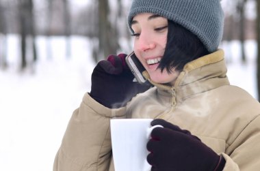 Oldukça çekici beyaz gözlü kız hipster kış Park portresi. Modern smartphone ve içecek kahve konuşuyor. Gençlik sokak moda. Açık havada yürüyüşler