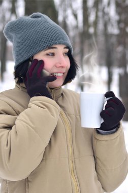 Oldukça çekici beyaz gözlü kız hipster kış Park portresi. Modern smartphone ve içecek kahve konuşuyor. Gençlik sokak moda. Açık havada yürüyüşler