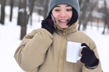Pretty genç kız sıcak kış şapkanı ve Paltonu giy. Soğuk havalarda smartphone ve kahve Kupası ile eğlenceli esmer kadın modeli yürümek dışında. Mutlu model poz gülümseyerek. İyi tatiller hava