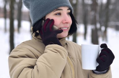 Oldukça çekici beyaz gözlü kız hipster kış Park portresi. Modern smartphone ve içecek kahve konuşuyor. Gençlik sokak moda. Açık havada yürüyüşler