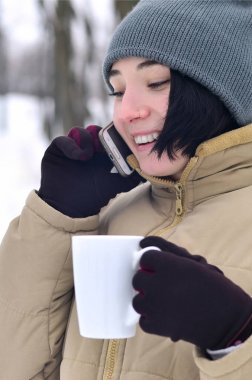 Oldukça çekici beyaz gözlü kız hipster kış Park portresi. Modern smartphone ve içecek kahve konuşuyor. Gençlik sokak moda. Açık havada yürüyüşler