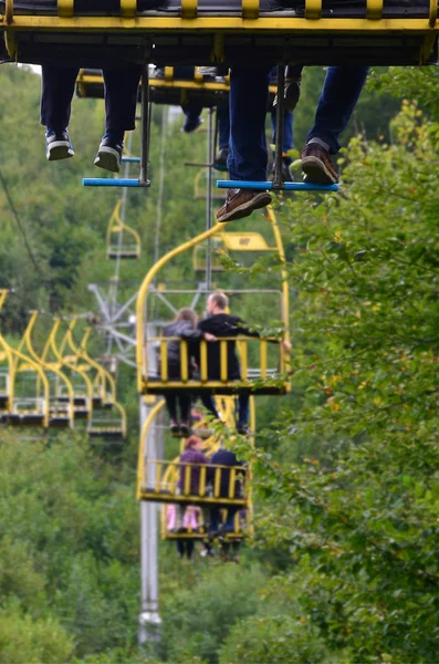 People ride on a cable car. The legs of passengers hang over the mountain forest