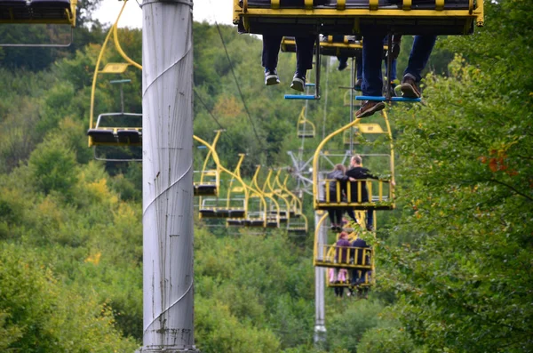 People ride on a cable car. The legs of passengers hang over the mountain forest