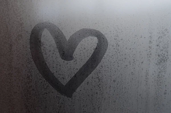 Love heart shape hand drawn on wet, frozen window pane with morning sunlight background. Selective focus macro picture