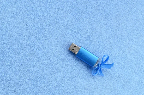 Brilliant blue usb flash memory card with a blue bow lies on a blanket of soft and furry light blue fleece fabric. Classic female gift design for a memory card