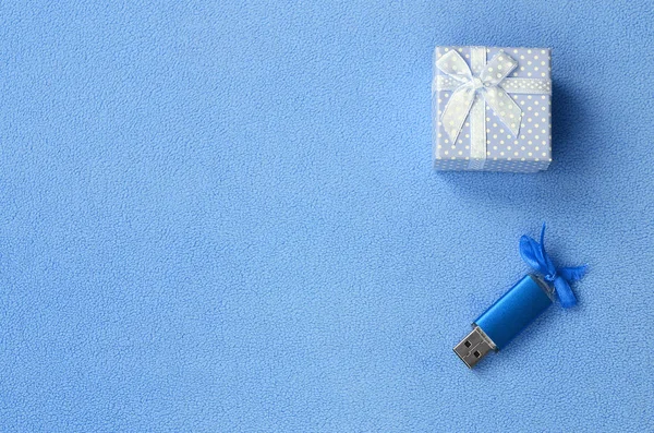 Brilliant blue usb memory card with a blue bow lies next to a small gift box in blue with a small bow on a blanket of soft and furry light blue fleece fabric. Classic female gift memory card design