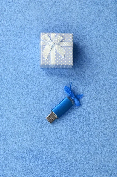 Brilliant blue usb memory card with a blue bow lies next to a small gift box in blue with a small bow on a blanket of soft and furry light blue fleece fabric. Classic female gift memory card design