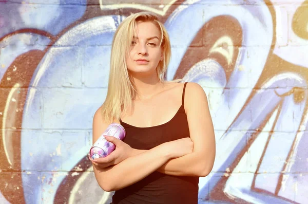 A young and beautiful sexy girl graffiti artist with a paint spray stands on the wall background with a graffiti pattern in blue and purple tones