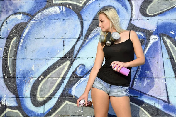 A young and beautiful sexy girl graffiti artist with a paint spray and gas mask on her neck stands on the wall background with a graffiti pattern in blue and purple tones